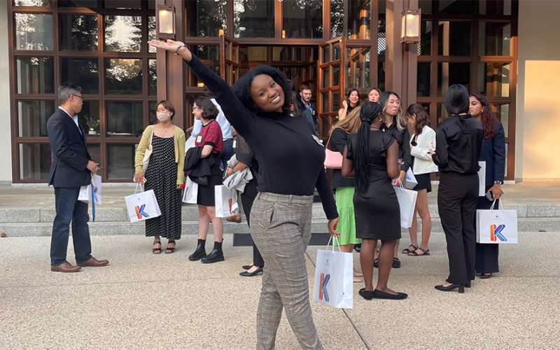 Alissa Banfield poses with a bag in one hand and an arm in the air outside of a building where a crowd has gathered.