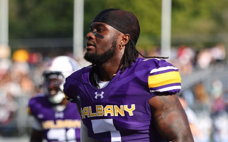 An African American man wears a bright purple jersey with the letters UALBANY across the front and the number 7. There is a football field, fans and other players in the background.