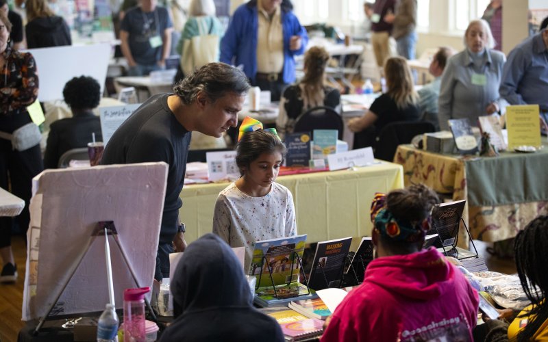 A man and a young girl look at a series of colorful, displayed books on tables as other people do the same in the background indoors.