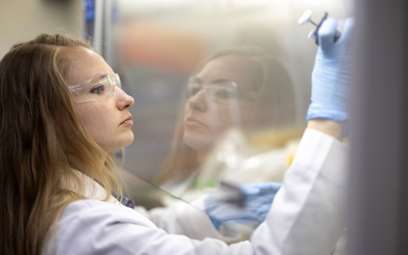 Tia Swenty, wearing safety glasses and gloves, works in a laboratory with a pipette inside a protective hood, her reflection visible on the glass