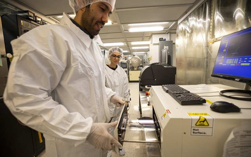 Two people in white cleanroom suits stand looking at a piece of white lab equipment in a semiconductor engineering lab.