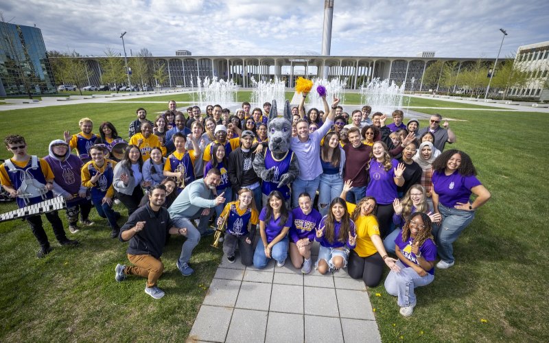 Students rally around The College Tour's host, Alex Boylan, in front of the Entry Plaza fountain. The group includes the cast and crew from the UAlbany episode of the Amazon Prime program as well as Damien, the Albany mascot, and UAlbany students.