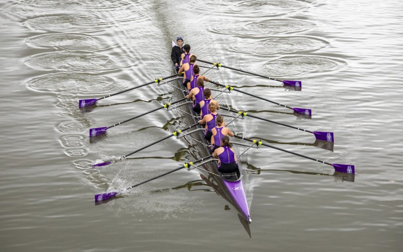 8 women and a coxswain from UAlbany row a long purple boat down a river as seen from an overhead shot