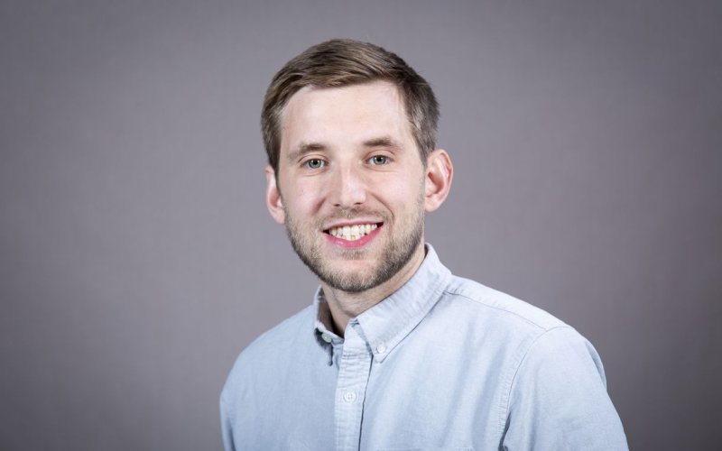 A man wearing a light blue button-down shirt smiles for a portrait against a gray backdrop.