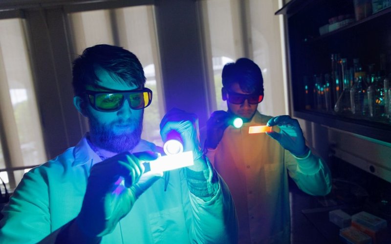 Two men in lab coats use handheld lights in a darkened lab to illuminate specimen slides that they are holding.