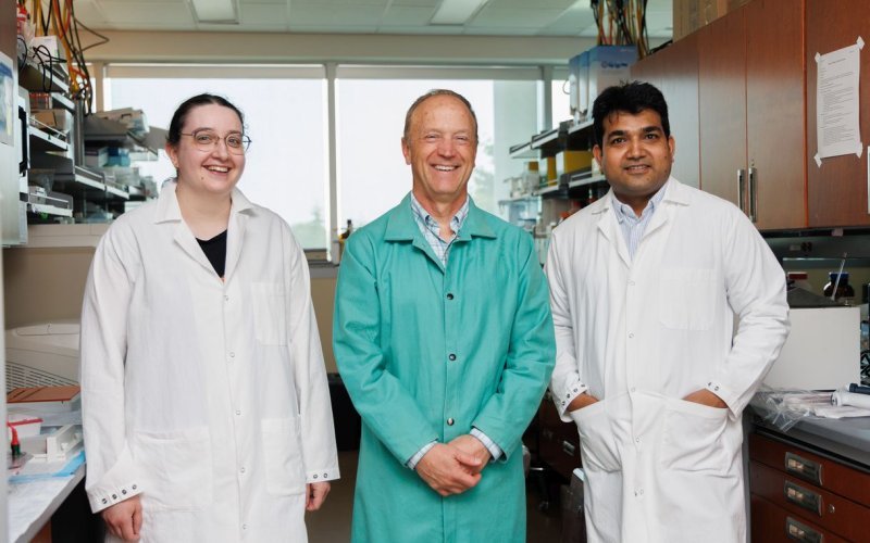 Three people, one woman and two men, stand together for a group portrait in a science lab. All are smiling and wearing lab coats. 
