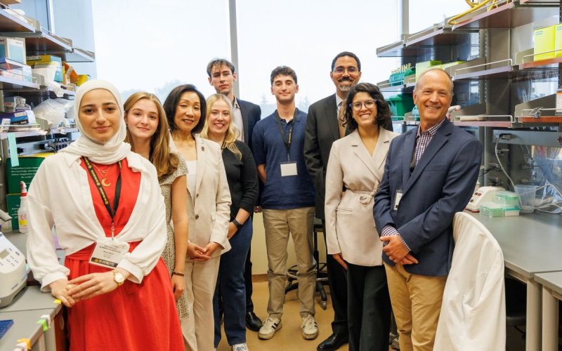 Nine smiling people stand together in a lab, posing for a group portrait. They are flanked by lab benches and shelving stocked with lab supplies.