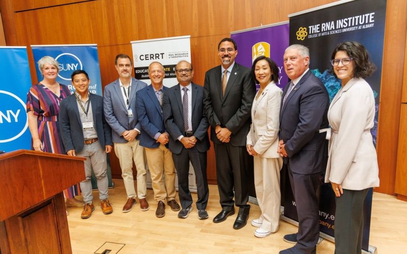 Nine people stand in a line behind a lectern smiling in front of banners for UAlbany, SUNY and the RNA Institute.