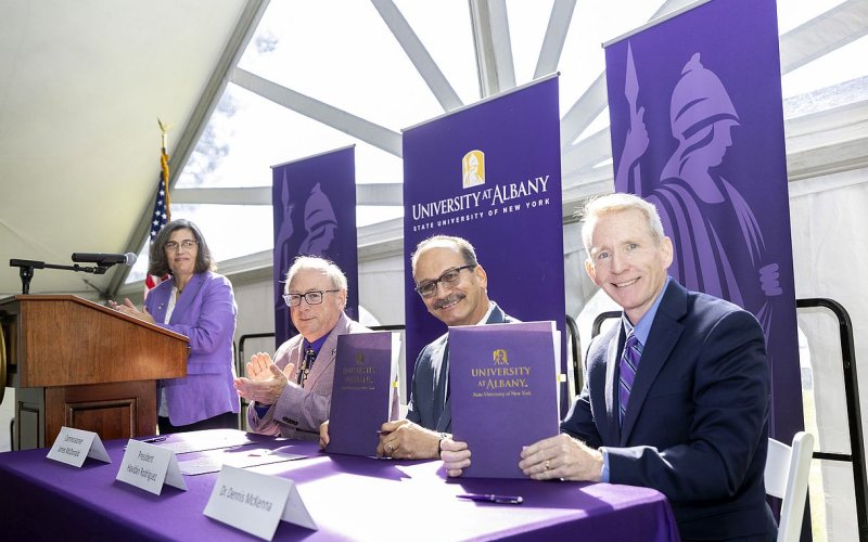 Four people are pictured under a white event tent with strong sunshine coming through. A woman wearing a purple blazer stands at a wooden podium. Three men wearing blazers sit at a table with a purple tablecloth, all smiling at the camera. Purple “University at Albany” banners are behind them.