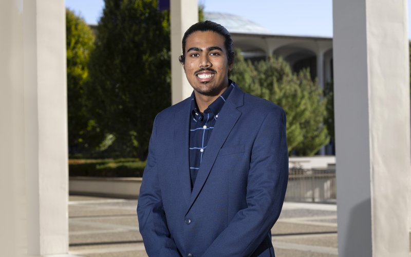 A young man stands outside amid a row of columns wearing a blue blazer and a blue shirt with trees and a dome in the background outside.