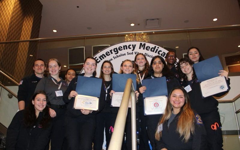 Smiling members of a student ambulance corps hold up certificates, posing on a staircase in front of a sign with the words "Emergency Medical" partially visible.