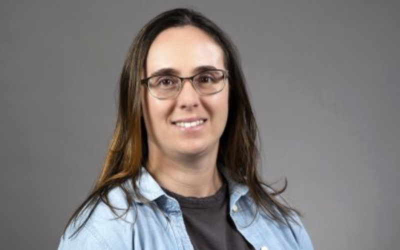 A woman wearing glasses and a denim shirt smiles at the camera