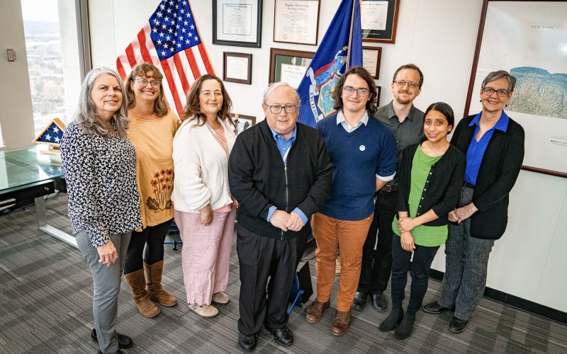 Eight people, including five women and three men, smile together for a group portrait in an office with gray carpet and a collection of framed plaques in the background. An American flag and the flag of New York State stand behind the group.