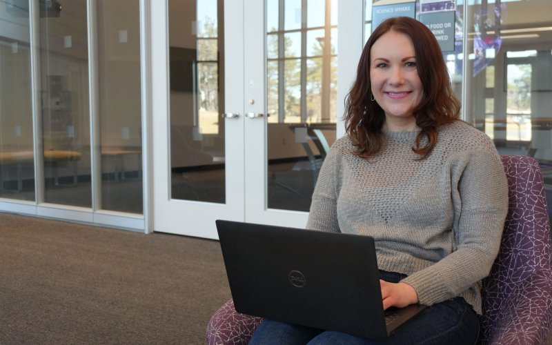 A woman with brown hair wearing a gray sweater and jeans sits in a purple chair, working on her laptop in UAlbany's ETEC building.