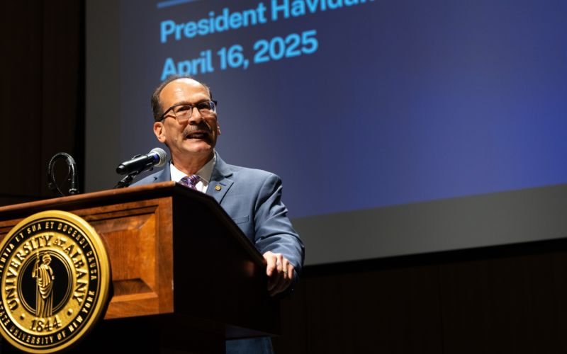 A man in glasses and a blue jacket speaks in front of a lecturn with the University at Albany seal.