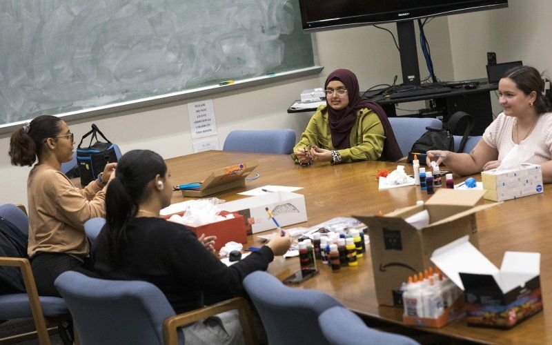 Four college students sit around a table strewn with bottles of paints and boxes, talking and doing crafts