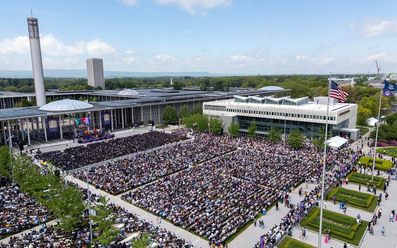 An aerial shot of the University at Albany main campus with a large crowd of people in the central plaza with a blue sky and large carillon in the background.