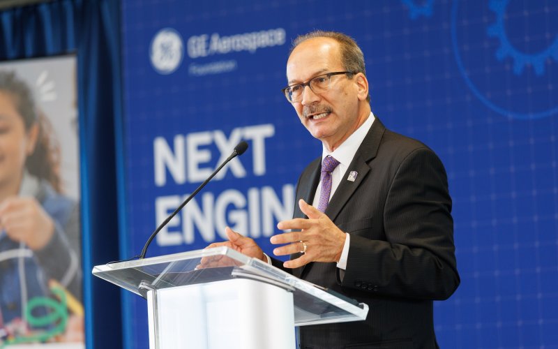 A man in a black suit, purple tie and glasses speaks at a podium on a stage in front of a screen that reads "Next Engineers"