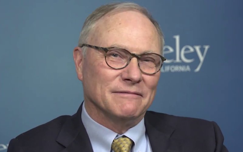 A man with balding hair and glasses, wearing a dark suit and yellow/gold checkered tie sits  for a portrait with a logo of the University of California, Berkeley behind him.