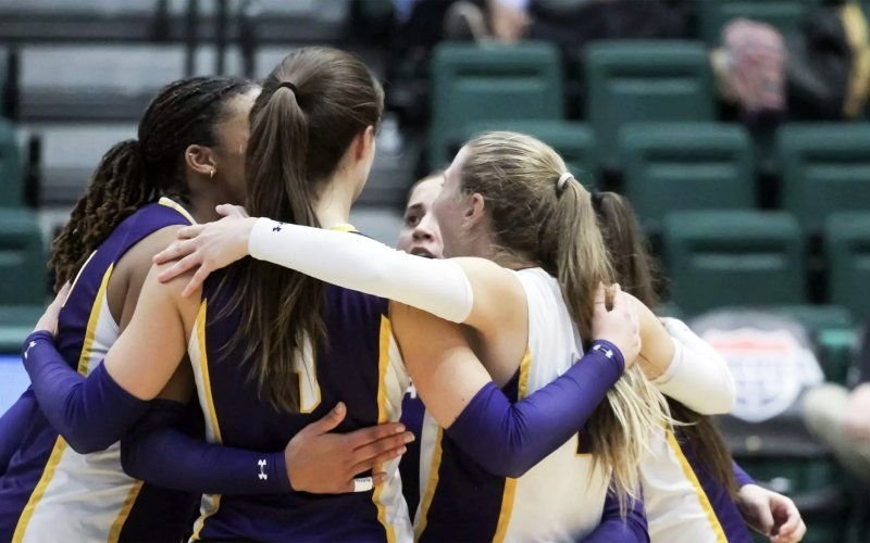 A group of women in purple, gold and white uniforms embrace each other in a huddle indoors with green chairs in the background.