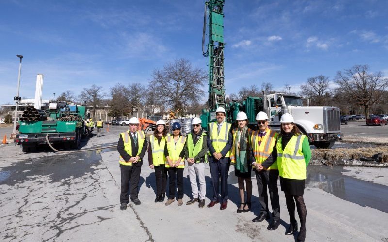 8 people, in green safety vests and white hardhats, stand in front of a tall green drill.