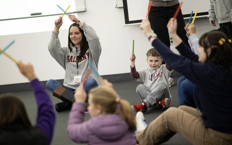 A woman in a UAlbany sweatshirt holds rhythm sticks over her head, sitting on the floor amid a group of children doing the same.