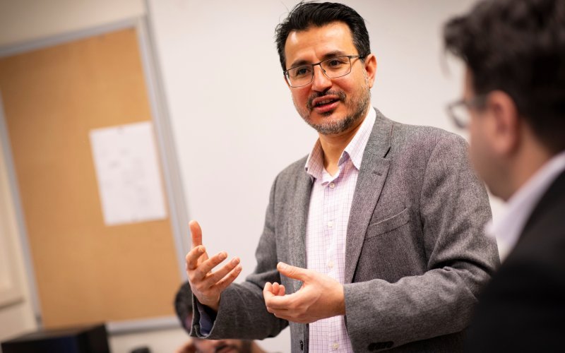 A man with dark hair and a gray blazer and glasses speaks animatedly in a brightly lit classroom. 