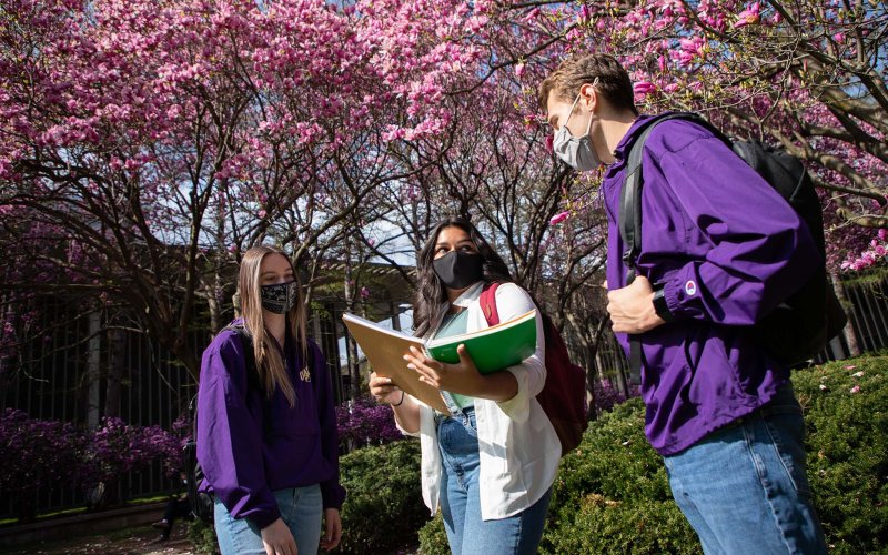 Three students, all masked, standing together talking outside. The student in the middle is holding an opened notebook.