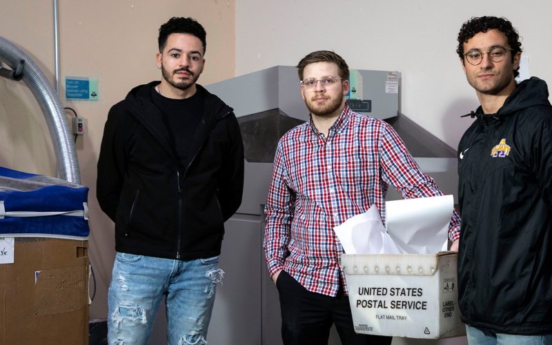 Three students stand indoors in front of a shredding machine and bypass that they created.
