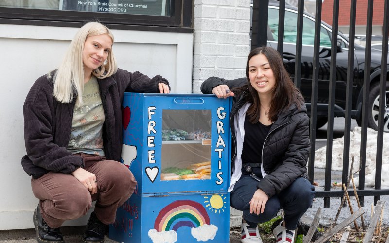Two smiling young women crouch on a sidewalk next to a blue metal box containing period products. A rainbow and the words “Free” and “Gratis” are painted on the front of the box. A glass window displays period products inside. 