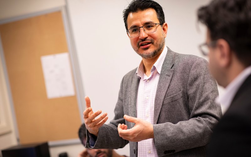A man with dark hair and a gray blazer and glasses speaks animatedly in a brightly lit classroom.
