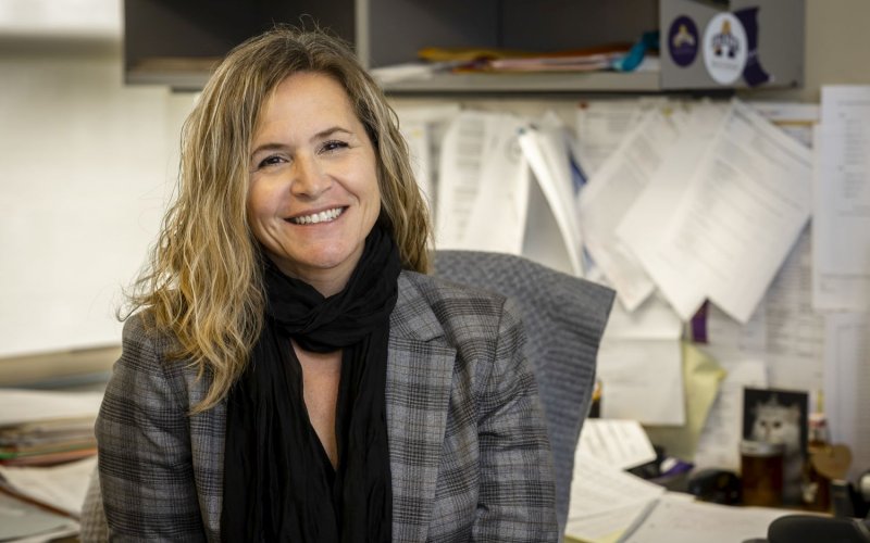 A smiling blond woman in a gray checked jacket and black scarf sits at a cluttered desk.
