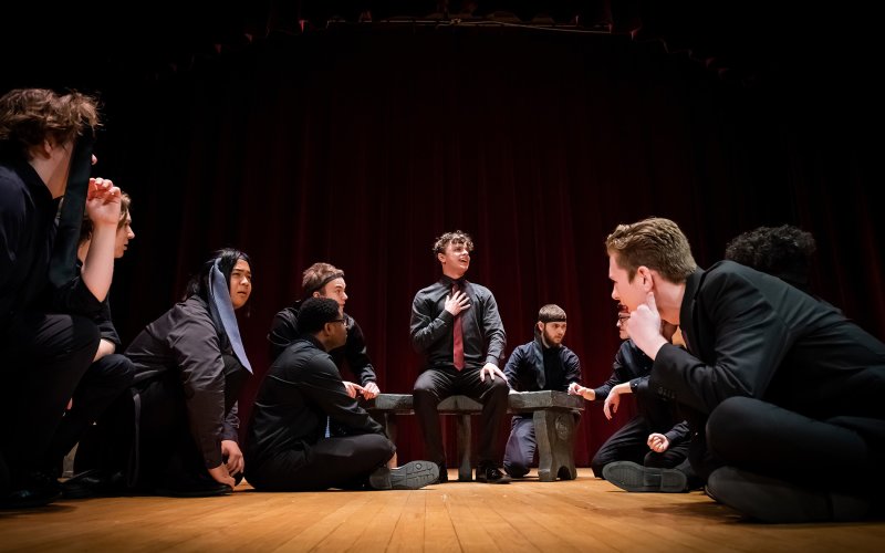 performers on floor dressed in black surround a singer seated on a bench