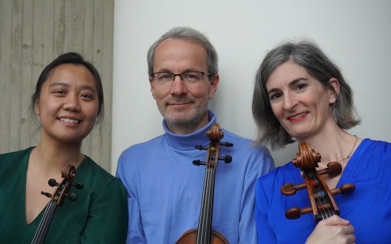 three smiling musicians stand in a line holding their instruments