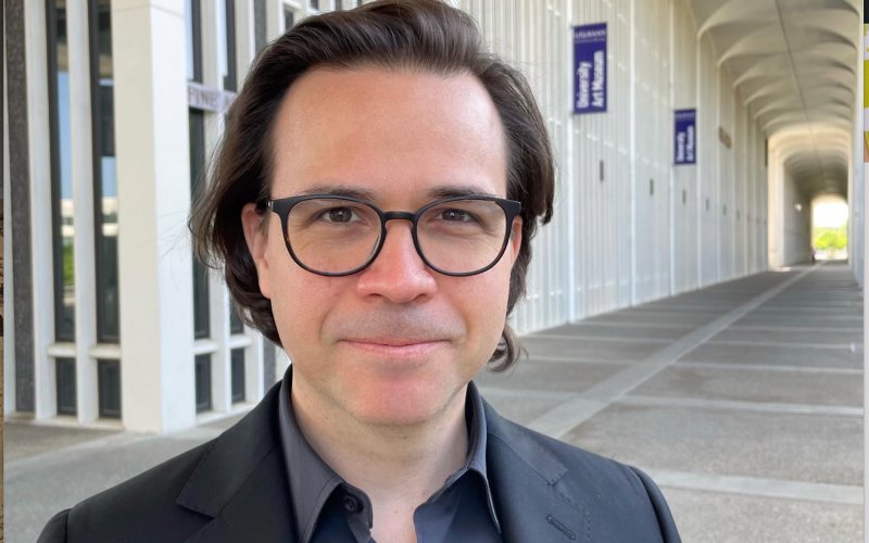 A man with short brown hair and black-rimmed glasses smiles outside of the University Art Museum at UAlbany on a sunny day.