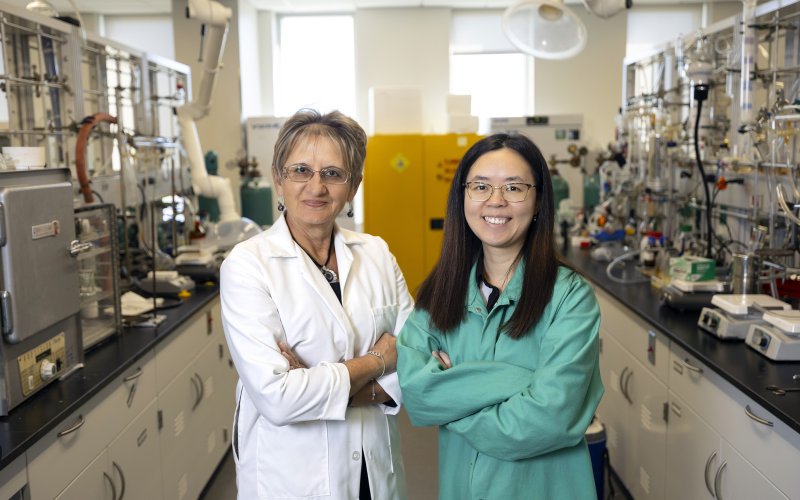 A older woman with short light brown hair and glasses in a white lab coat stands with a younger woman with long dark hair, glasses and a green lab coat in a chemistry laboratory with tools, beakers and chemicals.