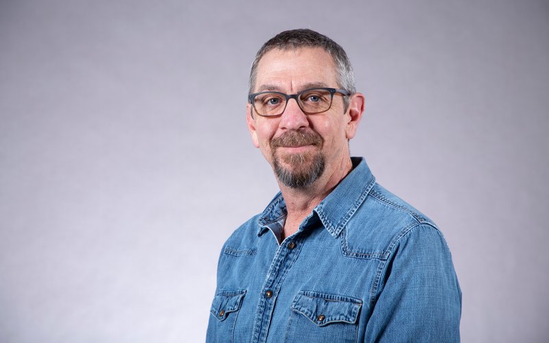 A man with short gray hair, a goatee and glasses wears a denim jacket and poses for a portrait against a gray backdrop.