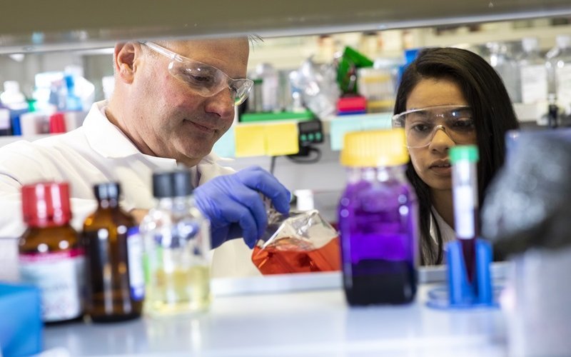 A photo of several glass bottles filled with colorful liquids with two scientists behind them in lab gear.
