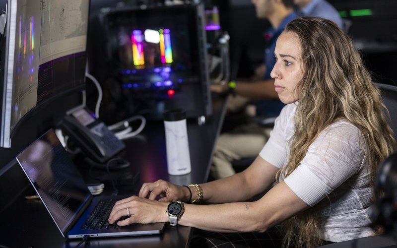 A woman in a white shirt sits at a laptop in a darkened computer lab with colorfully illuminated machines in the background. 