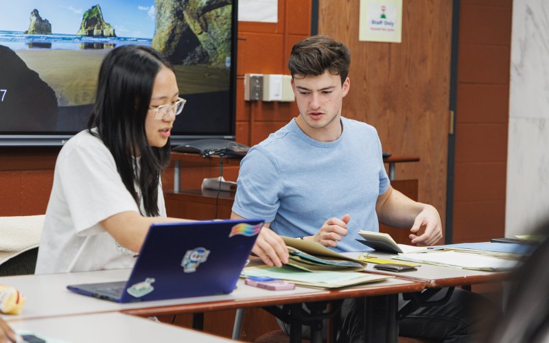 A young woman with dark hair, glasses and a white T-shirt sits next to a young man with brown hair and a blue t-shirt at a table as they go through folders of paper.
