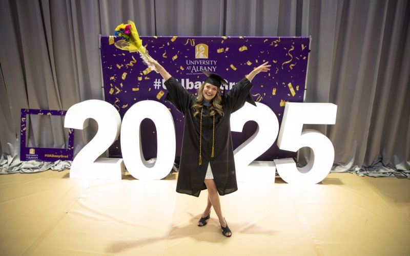 A UAlbany graduate student stands in cap and gown with arms upraised in front of a lit sign that reads 2025