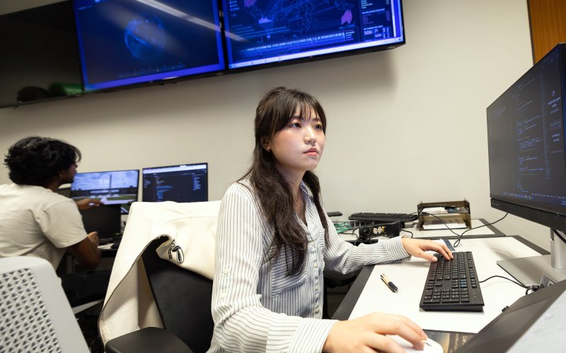 A female student with dark long hair and brown eyes wearing a striped blouse sits at a computer terminal with screens in the background displaying a digital globe in blue type.