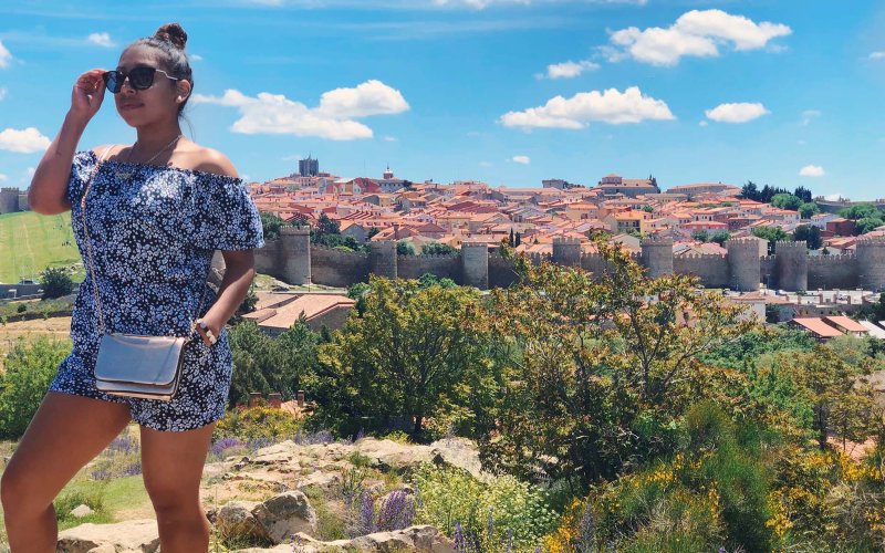 A young woman walks away from 100s of yards of castle walls and behind them a hillside of adobe-topped residences