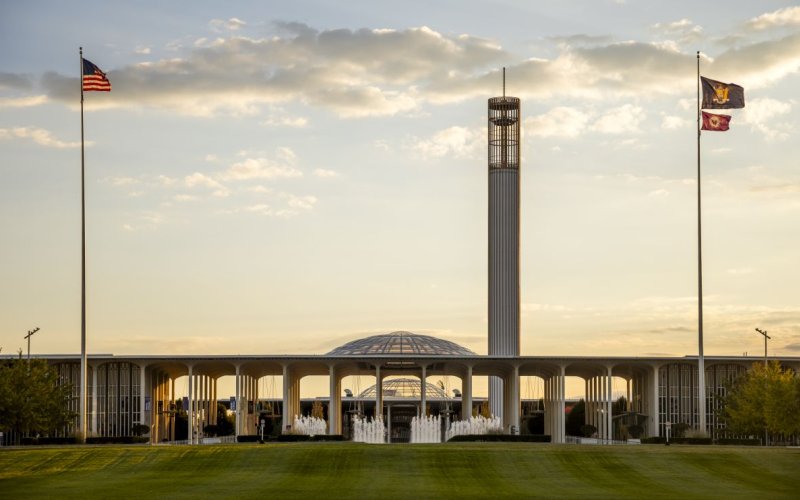 The UAlbany Academic Podium, Carillon and entrance fountain are seen at dusk under a darkening sky. Flags fly on poles on either side of the image.