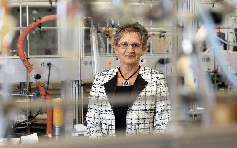 A woman with short hair, wearing glasses and a black and white tweed jacket with broad black lapels and bold accent jewelry, poses for a portrait in a chemistry lab. She is standing behind a lab bench hung with clear plastic tubing. Various tubes and glass lab containers are behind and around her.