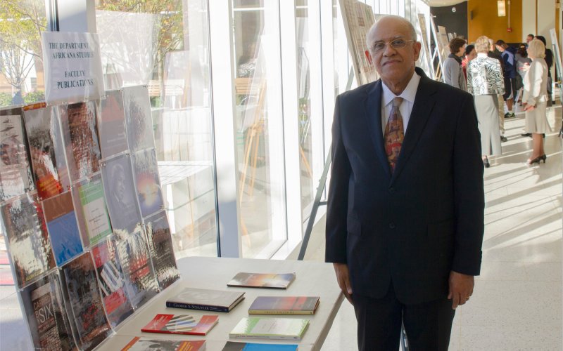 A man in a suit and tie and wearing glasses stands next to a table topped with books below a sign that reads "The Department of Africana Studies Faculty Publications"