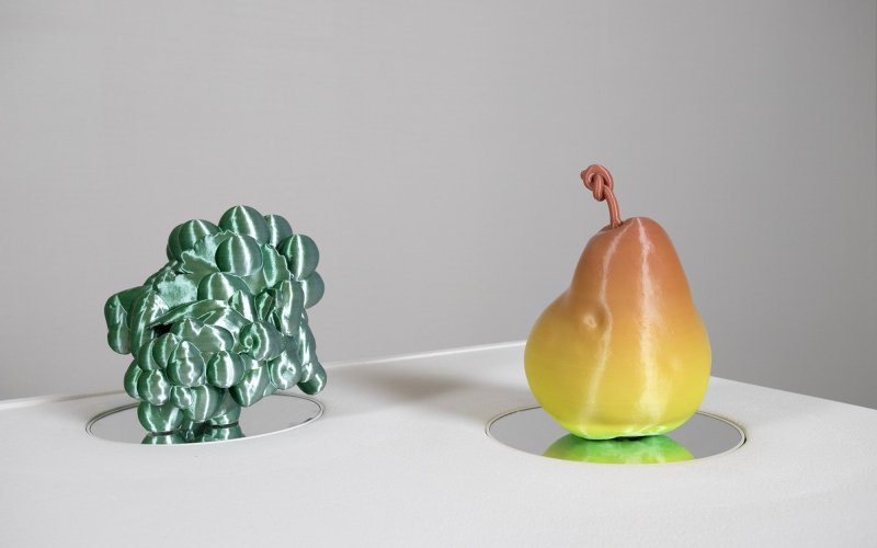 Some 3D printed grapes and a 3D printed pear sit on a table. The grapes are green and the pear is orange yellow and green.