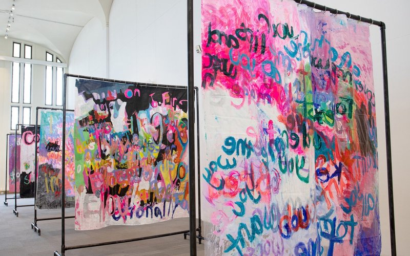 A series of frames stand in the exhibition space, colorful shapes and words painted onto canvasses hanging from them.