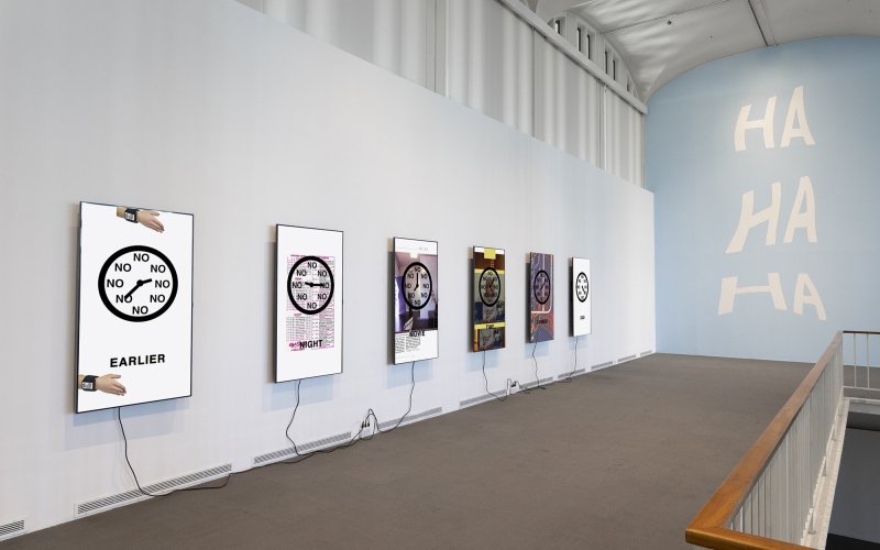 Six screens mounted on an exhibition wall, each playing variations of a clock with the word "no" replacing the numbers, on a wall farther away, "HA HA HA" is projected.