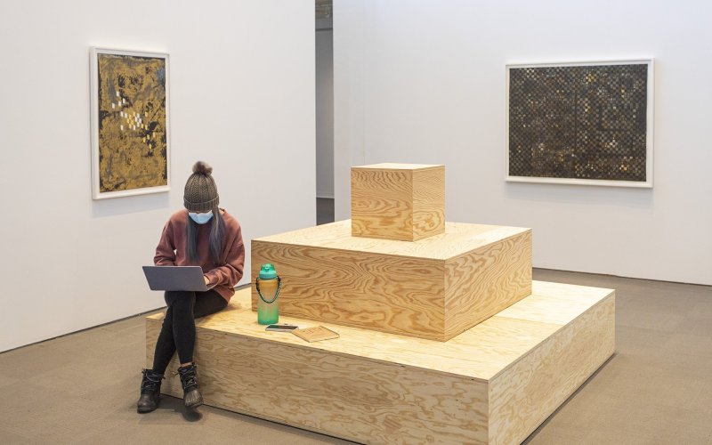 A person sits on a three-tired sculpture made from blocks of wood, using their laptop. On the walls in the space hang two art pieces.